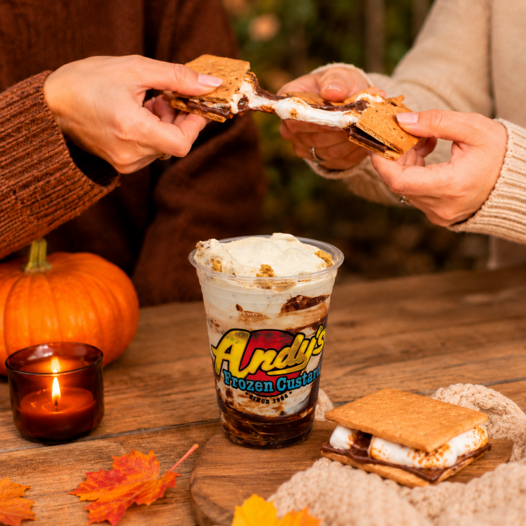 Two people pull apart a s’mores treat above a cup of Andy’s Frozen Custard on a wooden table with a pumpkin, candle, and autumn leaves.