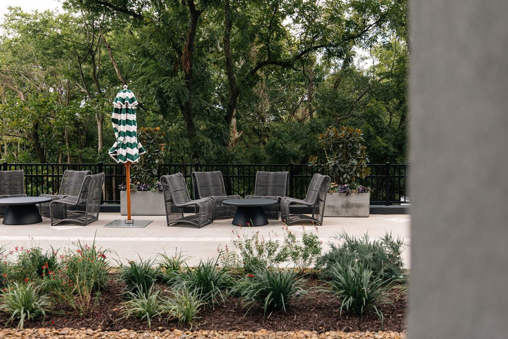 Outdoor patio seating area with wicker chairs and tables, a closed green and white striped umbrella, and a forested background.
