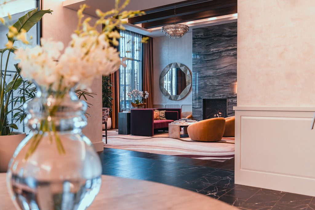 A hotel lobby lounge featuring a fireplace, a circular mirror, velvet seating, and a large crystal chandelier, viewed past a glass vase of flowers in the foreground.