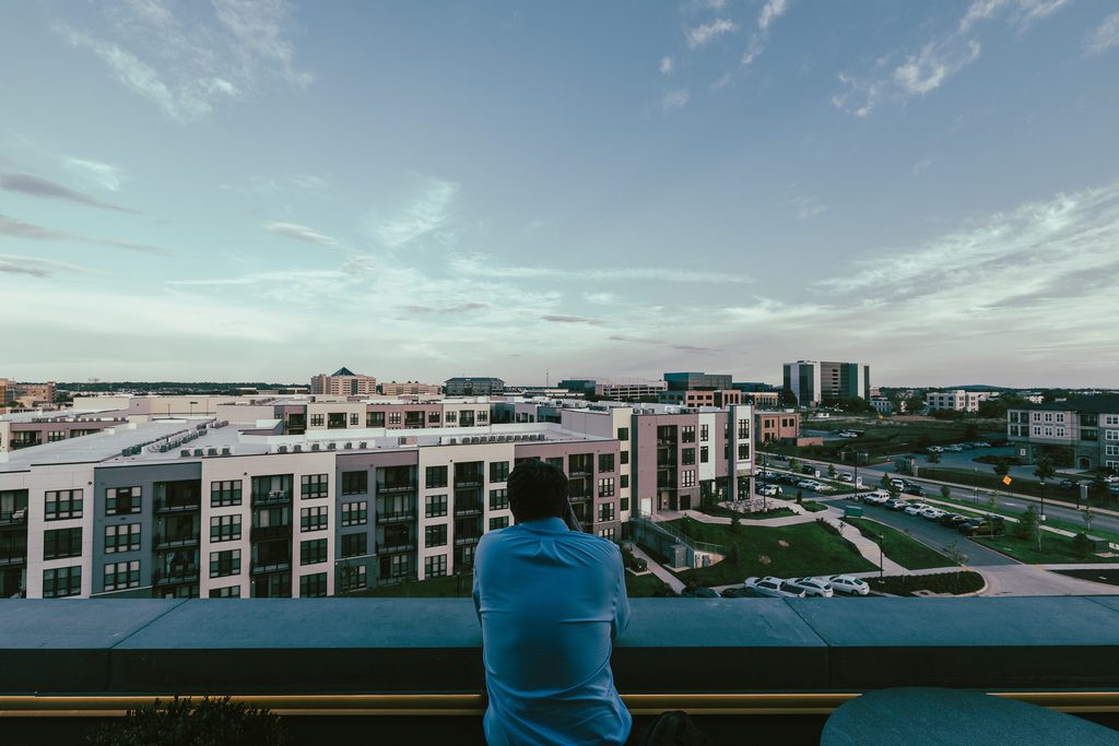 A person in a blue shirt looks out over an urban area with apartment buildings and a parking lot from a rooftop terrace.