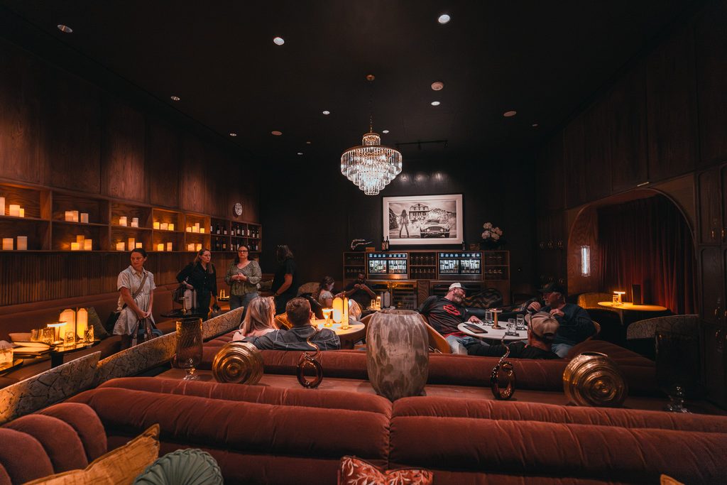 People sitting in a dimly lit, upscale lounge with dark wood paneling, velvet seating, and a large central chandelier.