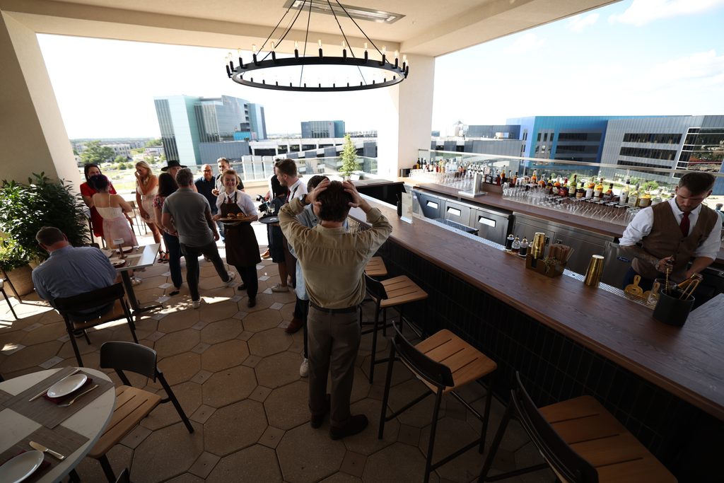 People gather at a rooftop bar with a bartender behind the counter and a server carrying a tray of drinks.