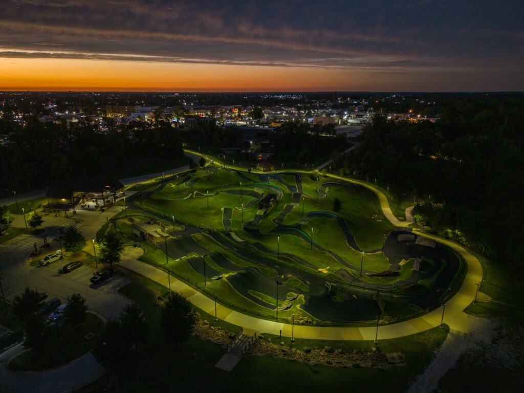Aerial view of a paved pump track illuminated at night, with a distant town skyline at sunset.